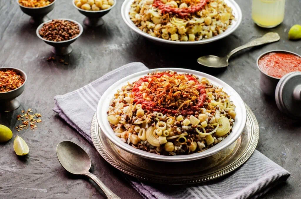 A bowl of Egyptian koshari layering rice, macaroni, brown lentils, and chickpeas, topped with crisp fried onions and a spiced tomato-garlic sauce, served on a guided Egypt all-inclusive tour.