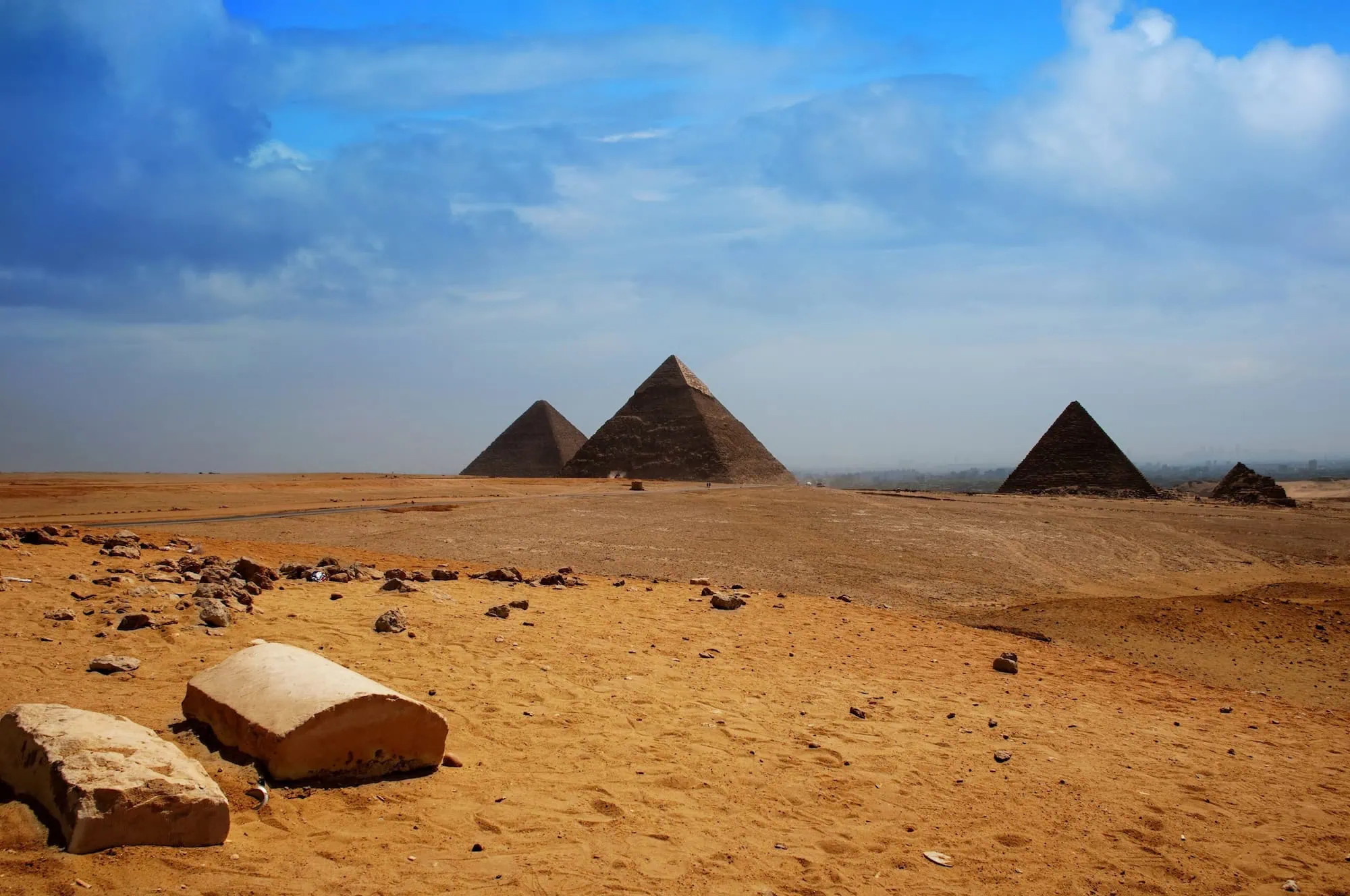The three Great Pyramids of Giza — Khufu, Khafre, and Menkaure — on the desert plateau west of Cairo, viewed across the golden sand foreground against a clear blue sky