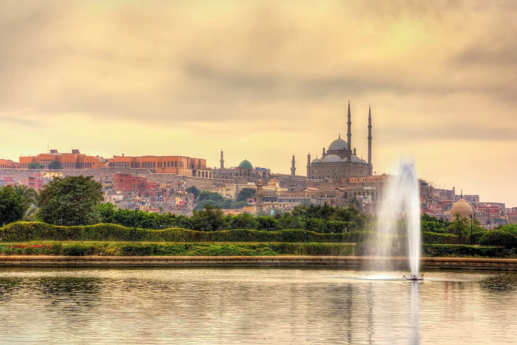 The Citadel of Saladin and Mohammed Ali Mosque with its distinctive central dome and twin minarets, viewed across a lake with fountains and gardens of Al-Azhar Park in Cairo at sunset.