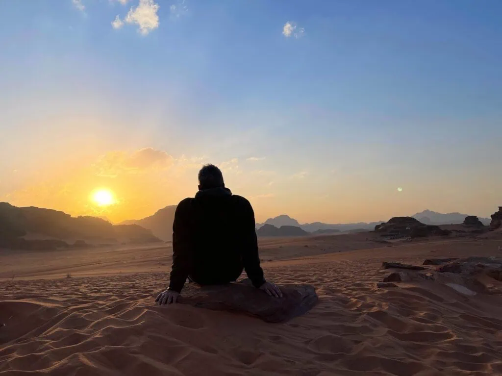 A traveller watching the sunset over the red sandstone mountains and desert sands of Wadi Rum, Jordan.