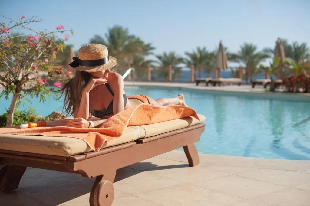 A traveler in a wide-brimmed straw hat reclining on a daybed beside a luxury hotel pool on Egypt's Red Sea coast