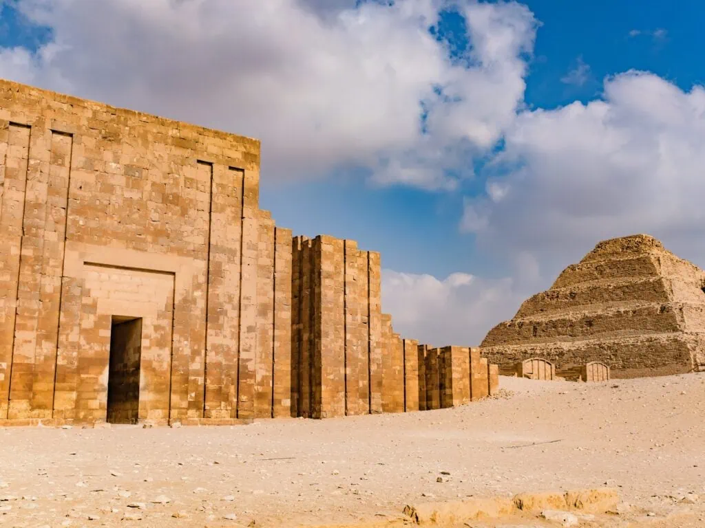 The Step Pyramid of Djoser at Saqqara visible in the background with its distinctive stepped form, and the bastioned limestone enclosure wall of the complex in the foreground, with the ancient entrance doorway visible in the wall.