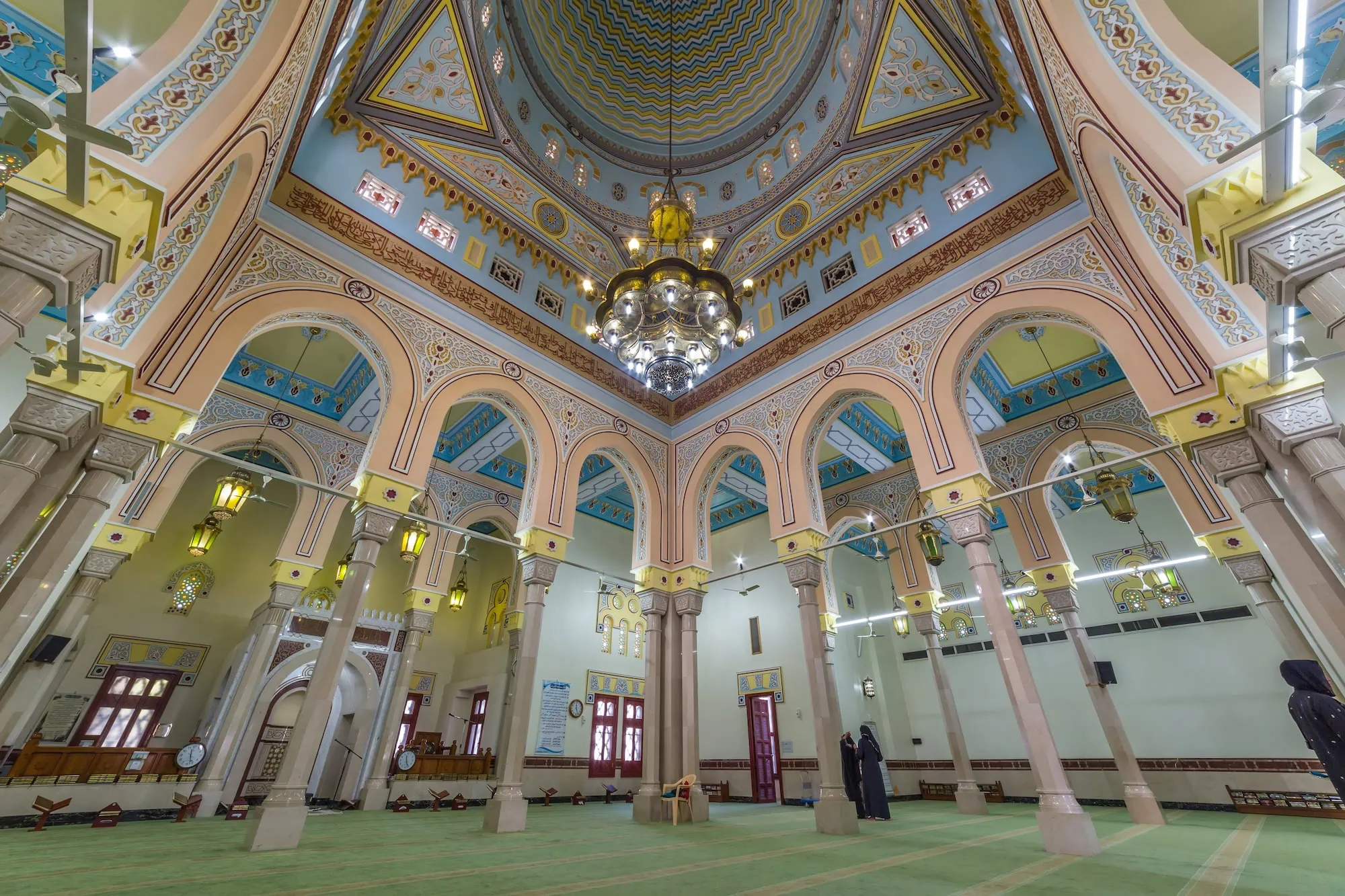 Interior of ornate mosque with decorated dome, chandelier, and visitors during guided tour