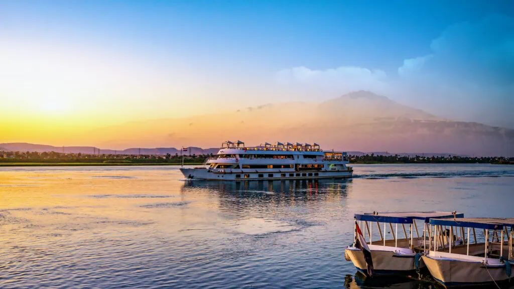 Nile River cruise ship sailing at sunset between Luxor and Aswan with mountains and palm trees