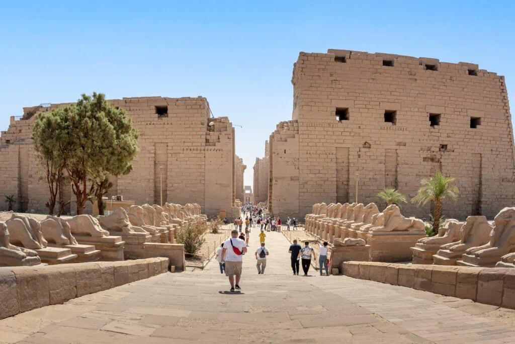 The massive sandstone First Pylon at Karnak Temple rising in the background, with a row of ram-headed sphinxes lining the processional avenue leading up to the entrance, photographed in warm light.