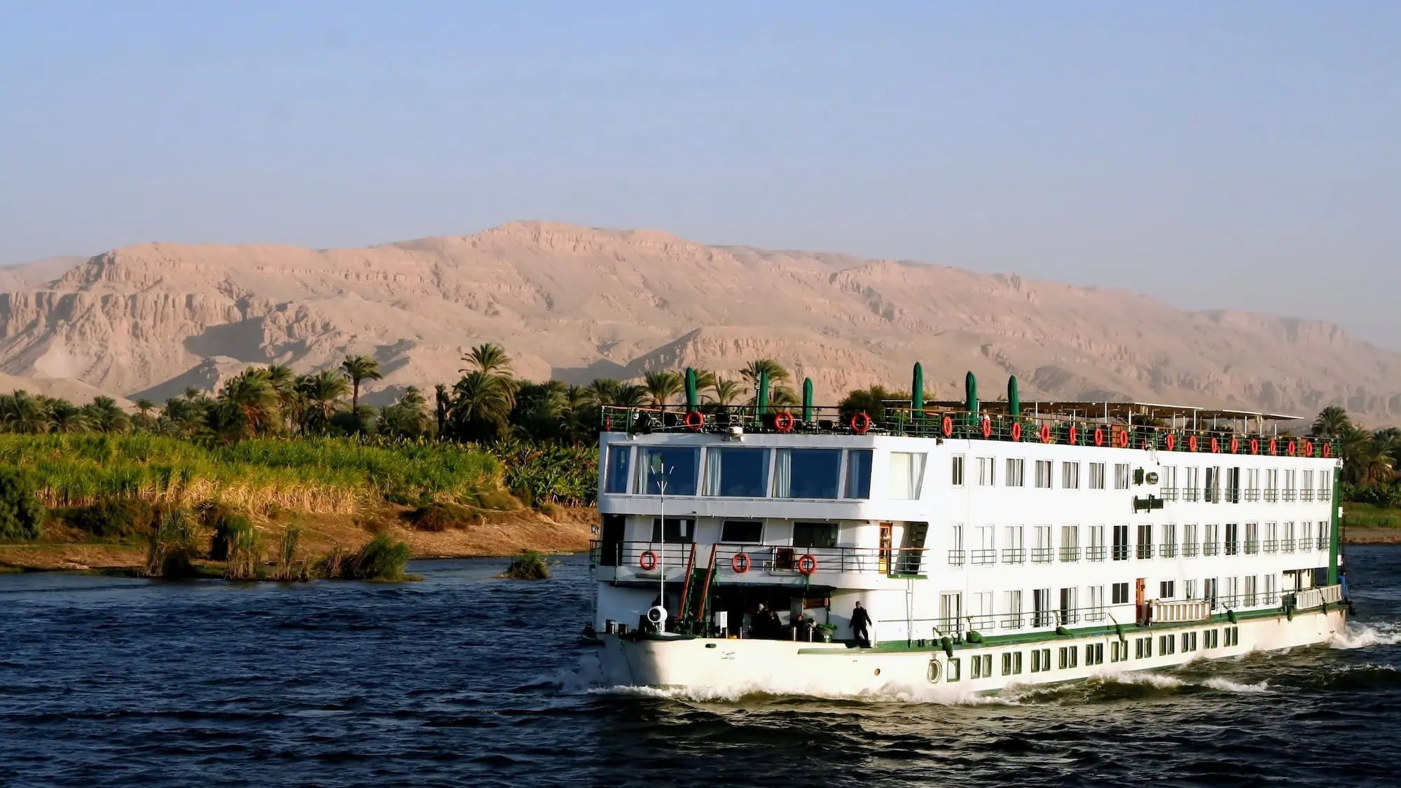 A multi-deck Luxury Nile Cruise vessel sailing the Nile River between Luxor and Aswan, Egypt — with desert hills and palm-lined banks in the background