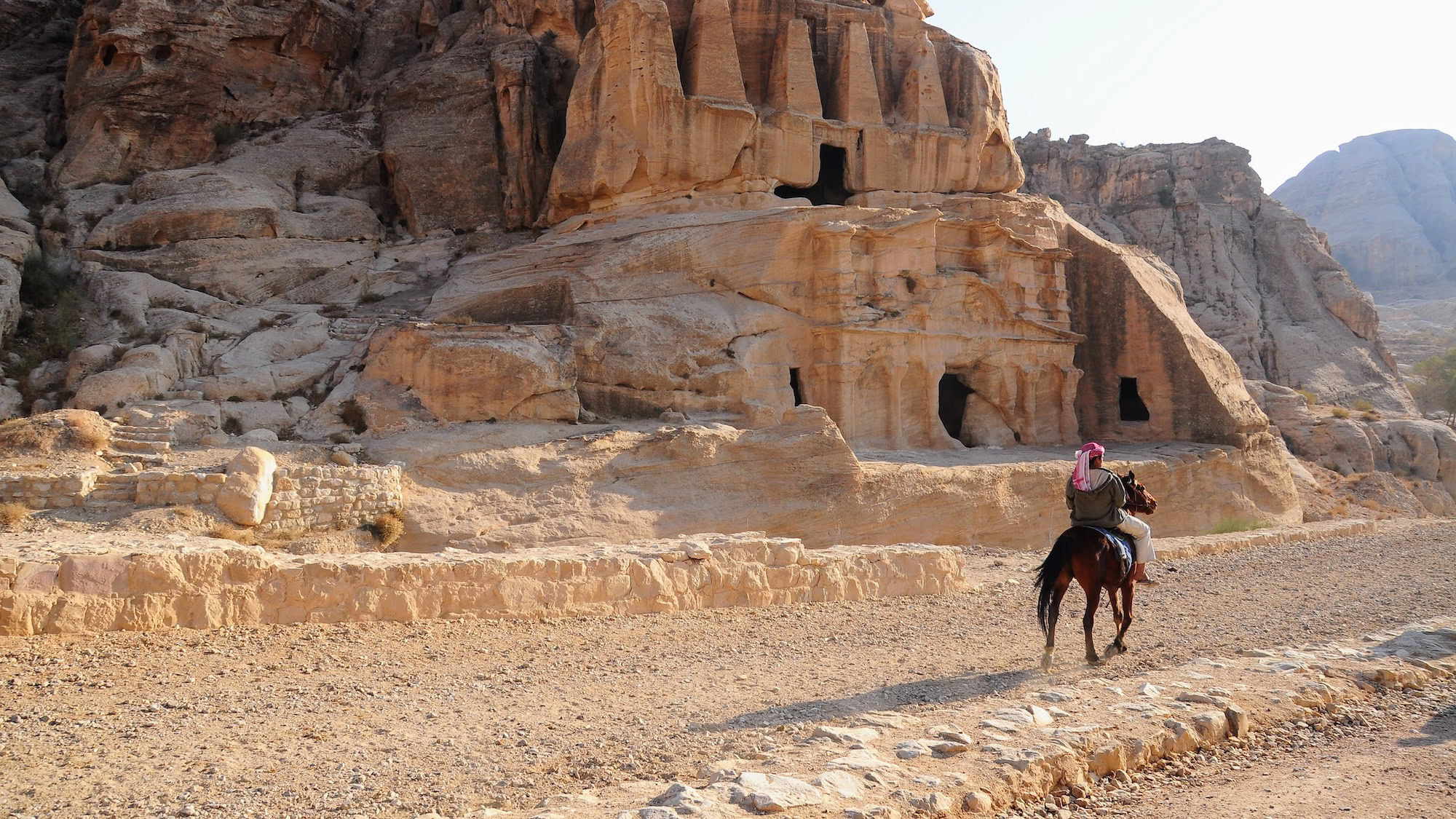 Person riding a horse through ancient rock-carved facades at Petra archaeological site