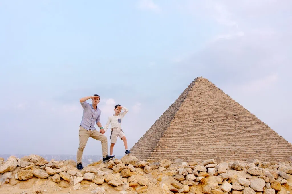 A father and young child standing together in light clothing on the desert plateau in front of the Pyramids of Giza