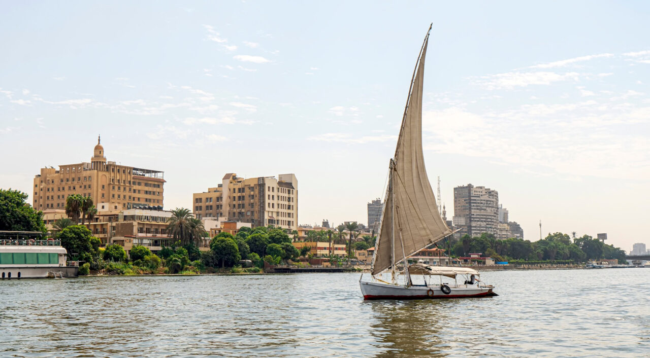A sailboat on a river walk sails along the Nile River in the center of Cairo among the skyscrapers and attractions.