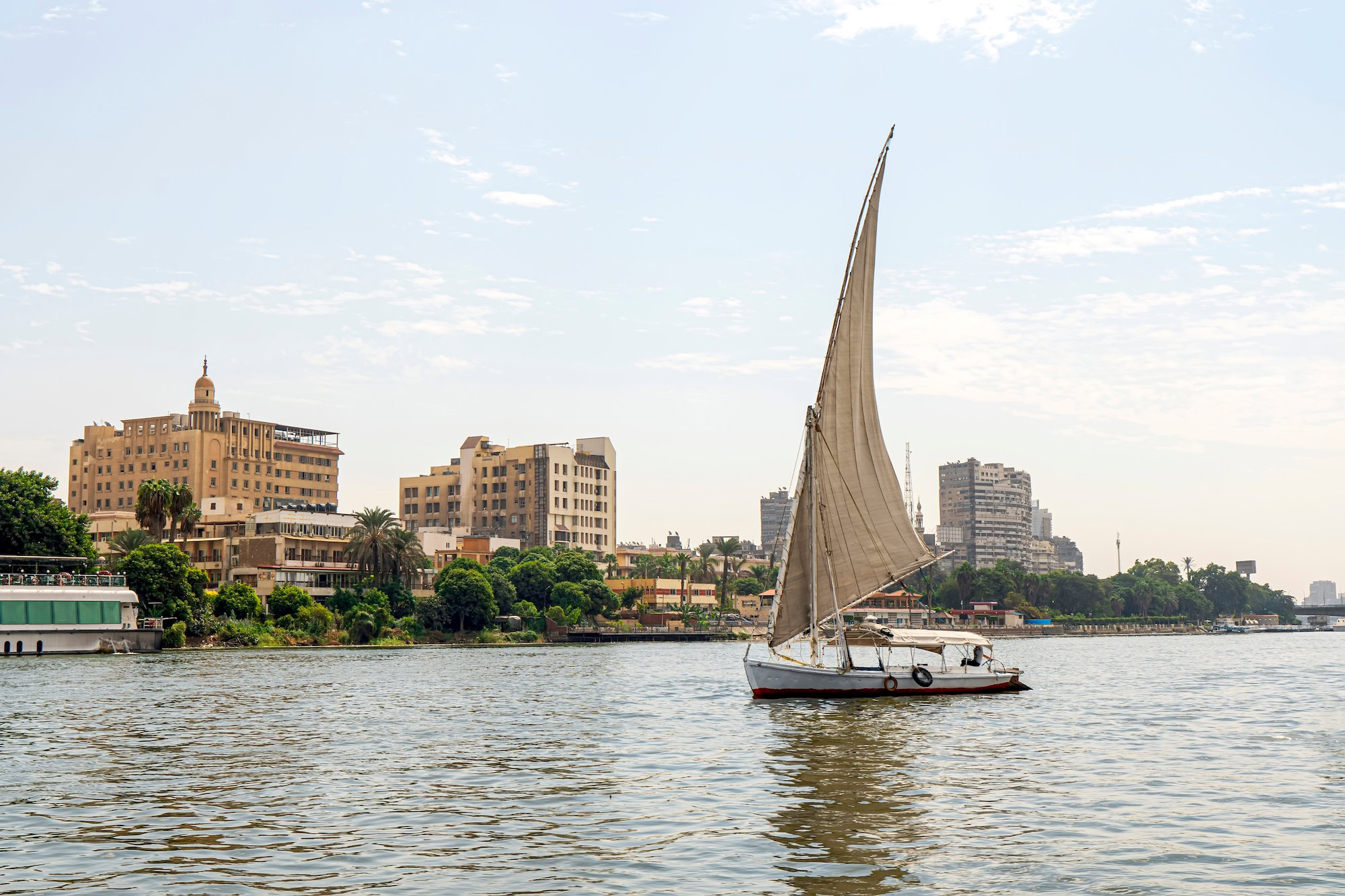A sailboat on a river walk sails along the Nile River in the center of Cairo among the skyscrapers and attractions.