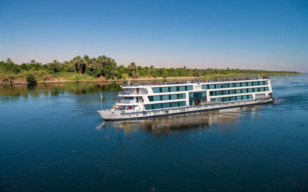 A multi-deck Luxury Nile Cruise vessel sailing past palm-lined banks on the Nile in Upper Egypt — 5-star vessel with panoramic cabins, sailing the standard Luxor–Aswan route over 4 days
