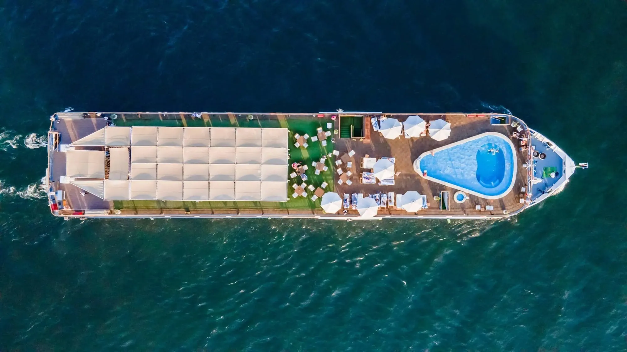 Aerial view of the upper sun deck of a 5-star Luxury Nile Cruise vessel sailing the Nile between Luxor and Aswan, with swimming pool, loungers, and bar area, typical of the 50- to 150-guest vessels operating the standard 4-day cruise itinerary and longer 7-night options.