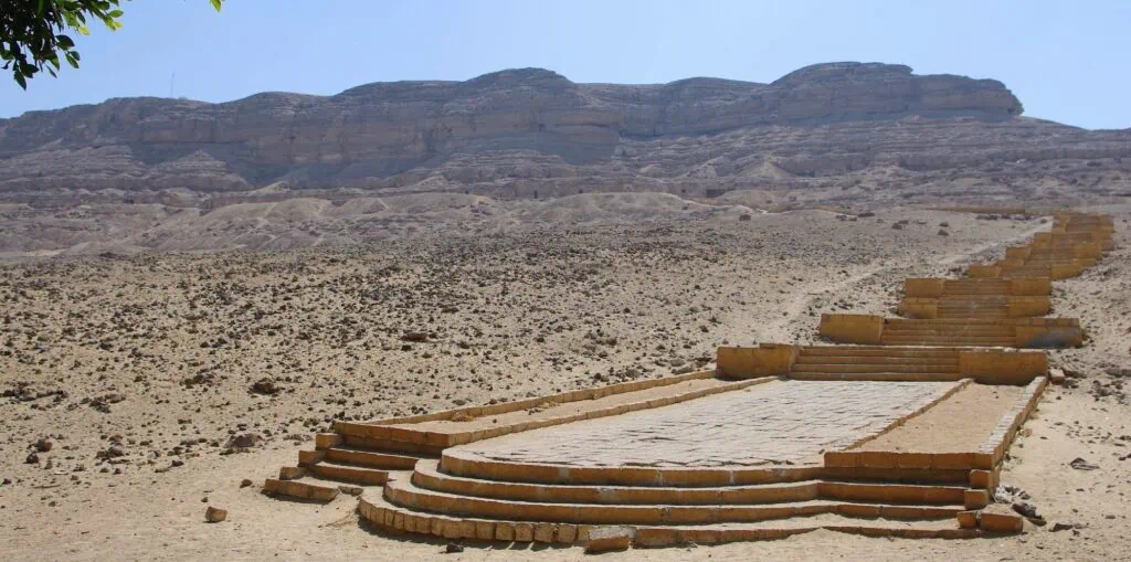 The Beni Hassan archaeological site in Middle Egypt, showing the limestone cliff face containing the rock-cut tombs, with a modern stone stairway and access platform leading up from the desert below.