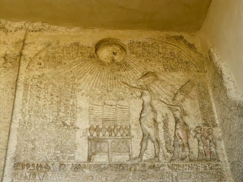 An ancient Egyptian boundary stela carved into a cliff face, showing the sun disc Aten with rays ending in hands, Akhenaten and Nefertiti making offerings at an altar, with hieroglyphic inscriptions covering the surrounding surface.