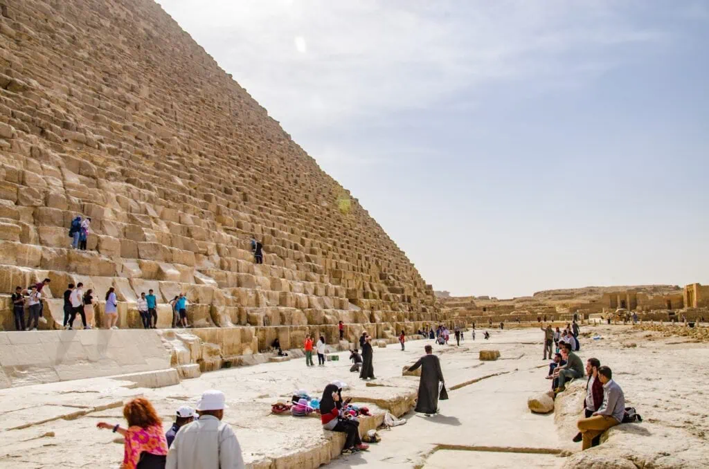 Visitors standing at the base of the Great Pyramid of Khufu (Cheops) on the Giza plateau, dwarfed by the massive limestone blocks, Cairo, Egypt. 7-day Egypt tours.