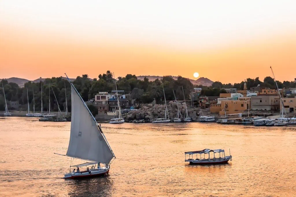 A traditional felucca sailboat on the Nile River at sunset near Aswan, with a colourful Nubian village visible on the riverbank in the background, Egypt.