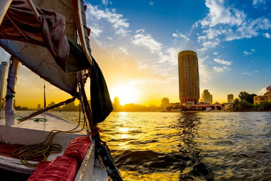 A traditional felucca sailboat with a white sail gliding on the Nile River at sunset, with the Cairo city skyline silhouetted in the background, Egypt.