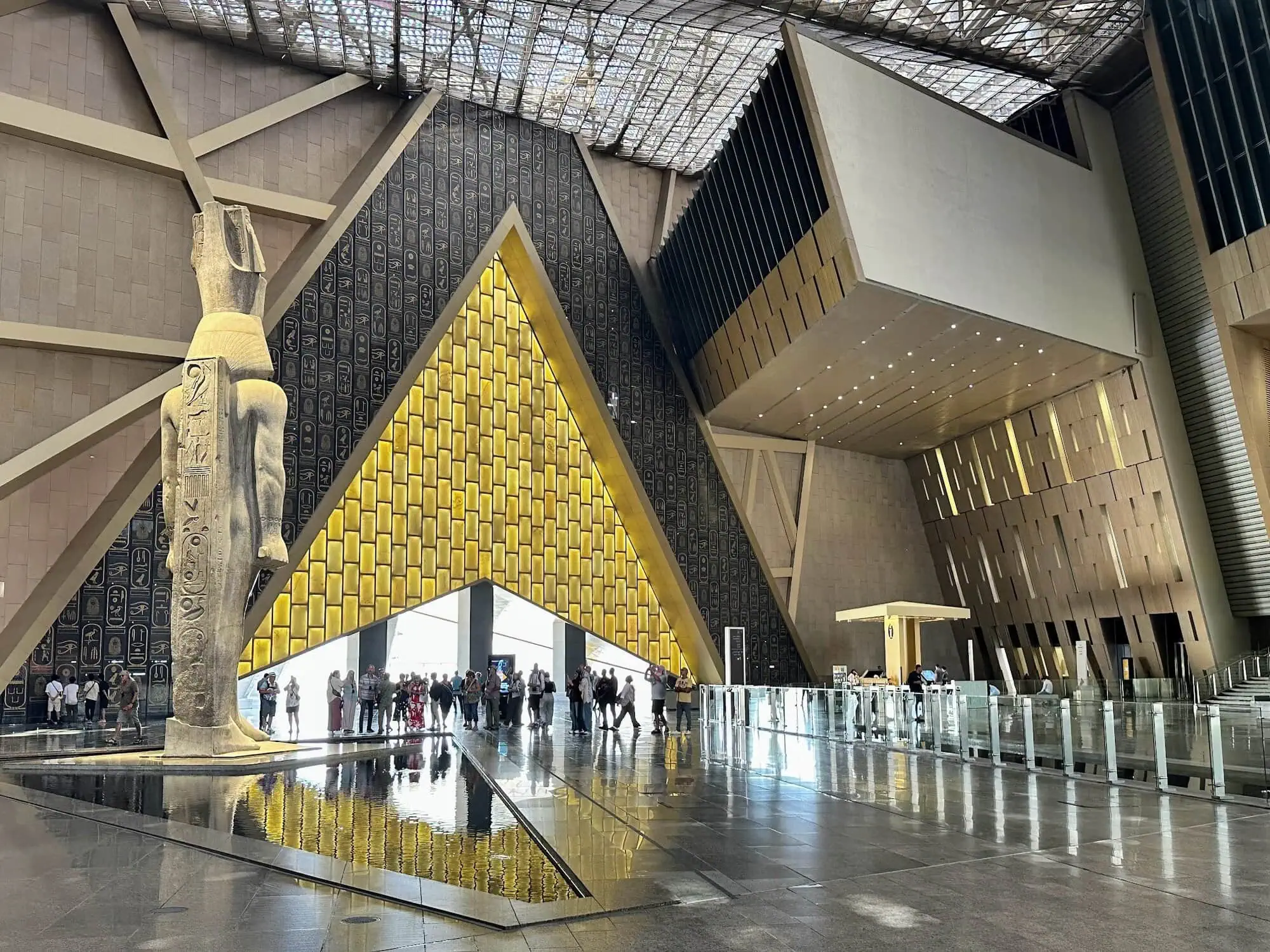 Interior of Grand Egyptian Museum showing modern architecture with glass ceiling, hieroglyphs, and visitors