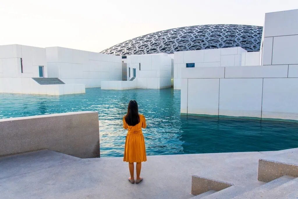 A visitor beneath the perforated dome of the Louvre Abu Dhabi on Saadiyat Island, UAE — the museum opened in 2017, designed by Pritzker Prize-winning architect Jean Nouvel, with daylight filtering through the eight-layered metal canopy