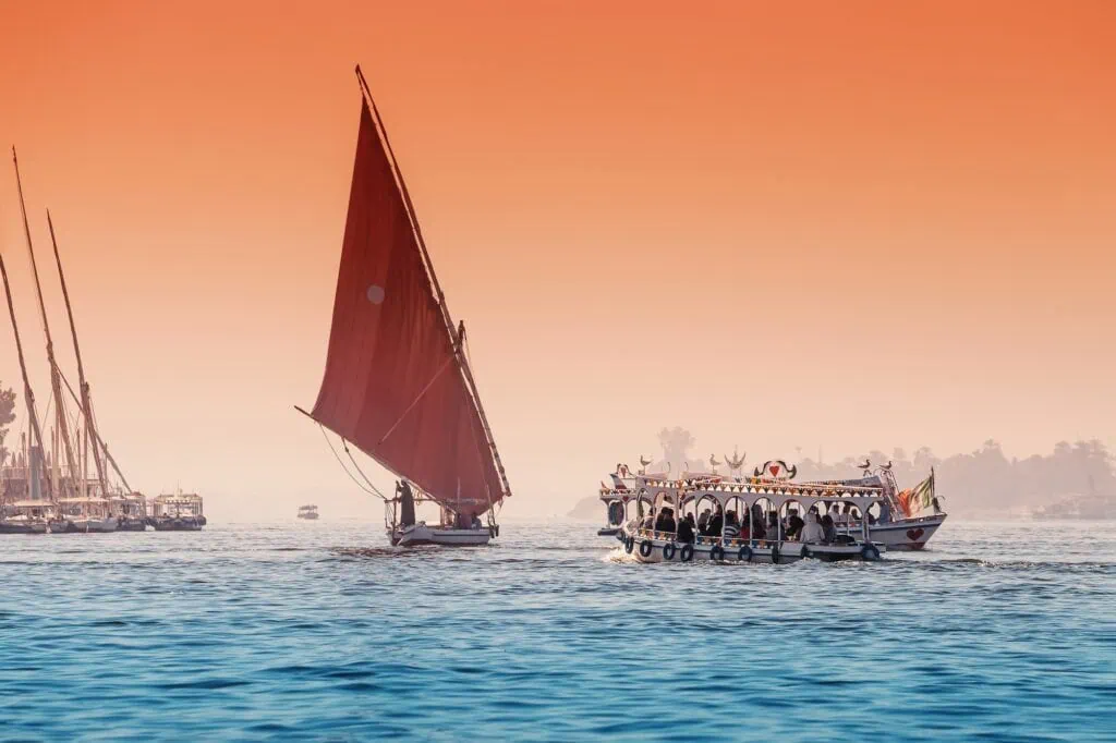 A traditional felucca sailboat with a orange lateen sail gliding along the Nile River past the banks near Aswan, Egypt.