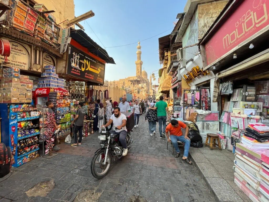 A busy street view in the Khan El Khalili bazaar in Islamic Cairo, with local shops, pedestrians, and a mosque minaret rising in the background, Egypt.