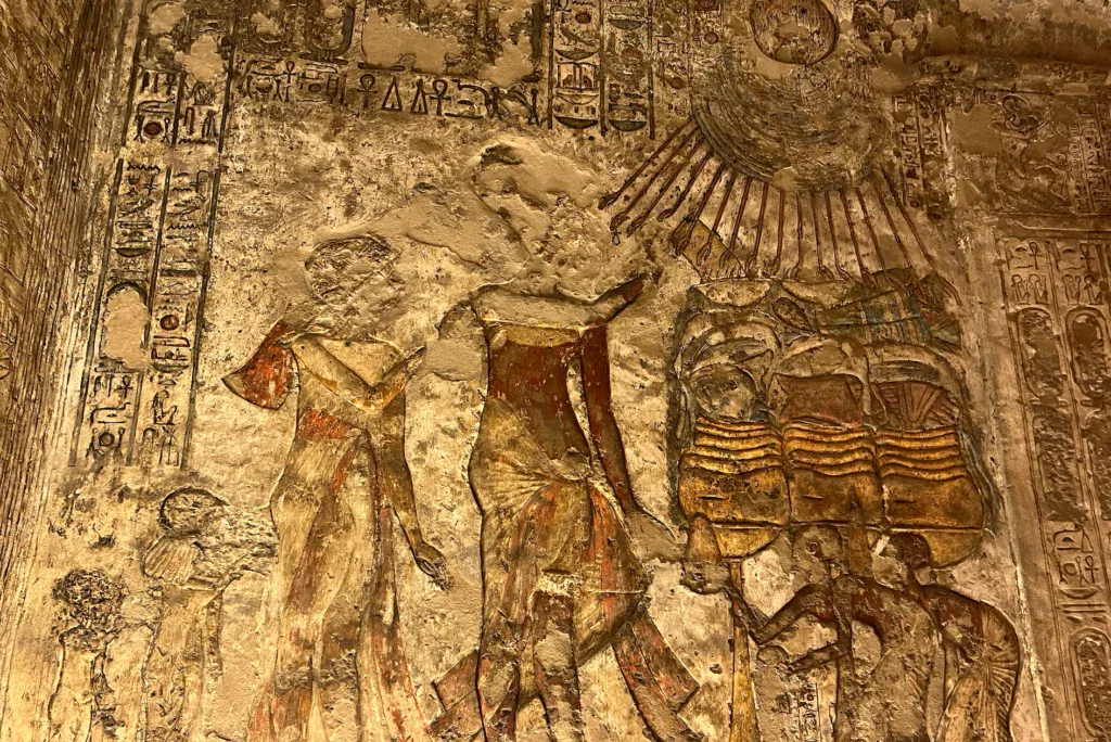 An ancient Egyptian limestone relief from Tell el-Amarna showing Pharaoh Akhenaten and his family standing beneath the sun disc Aten, whose rays descend toward them ending in small hands.
