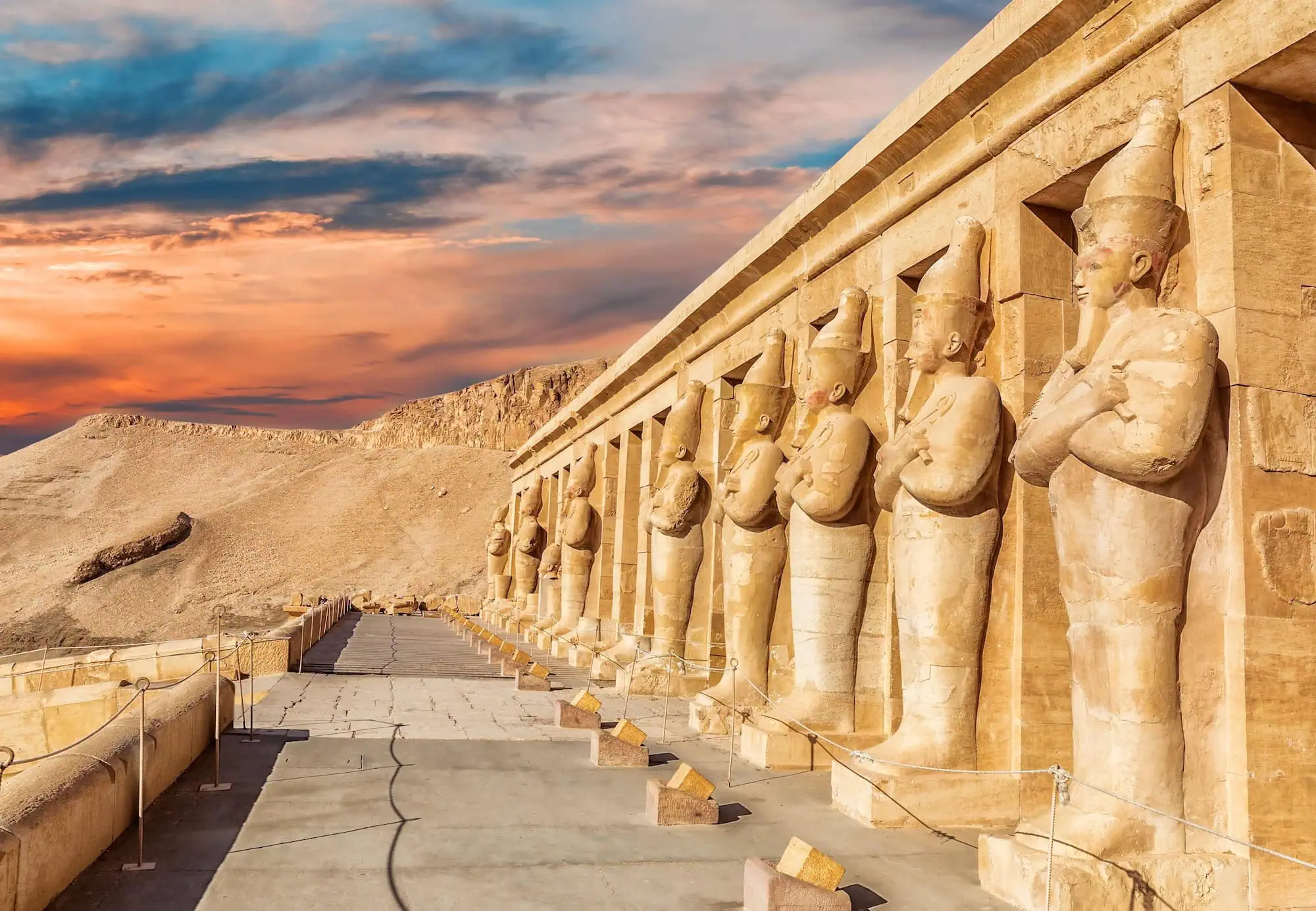 Ancient Egyptian pharaoh statues and limestone columns at Hatshepsut's mortuary temple against dramatic desert cliffs