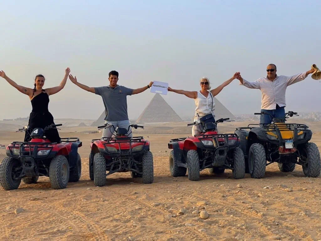 A group of travelers on quad bikes riding across the desert plateau with the Pyramids of Giza visible in the background