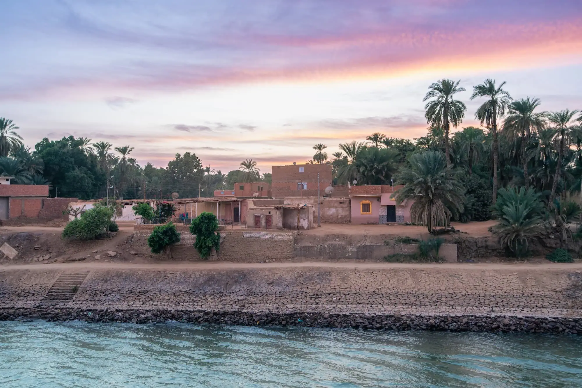 A traditional Egyptian village along the banks of the Nile River at sunset, with palm trees, mud-brick houses, and the river glowing in the warm evening light.