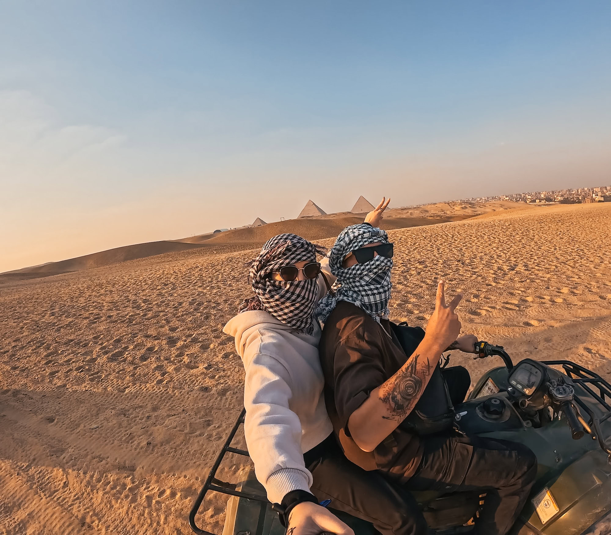 Couple taking selfie while riding quad bike and making peace sign with giza pyramids in background during sunset