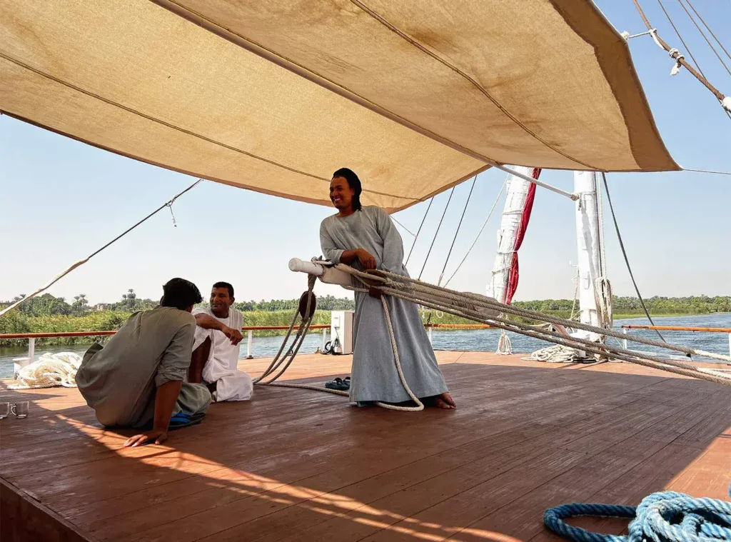 A dahabiya crew member preparing the sun deck of a traditional Egyptian sailing vessel on the Nile, with table settings ready for afternoon service