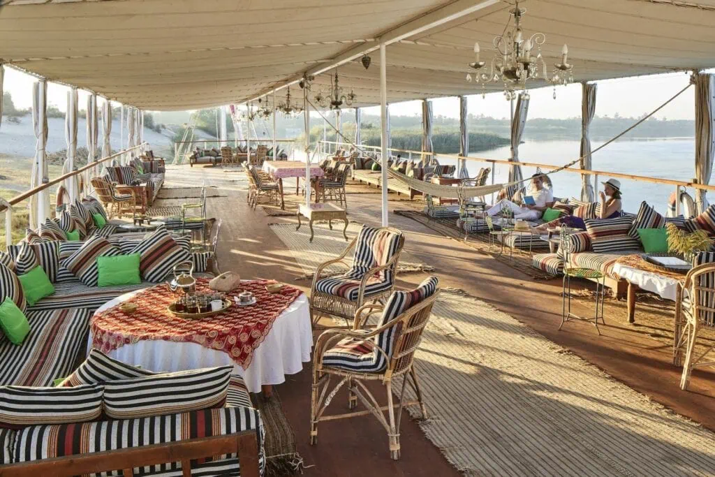 Travelers reading and relaxing on the wooden sun deck of an Egyptian dahabiya with the Nile River and palm-fringed banks visible in the background