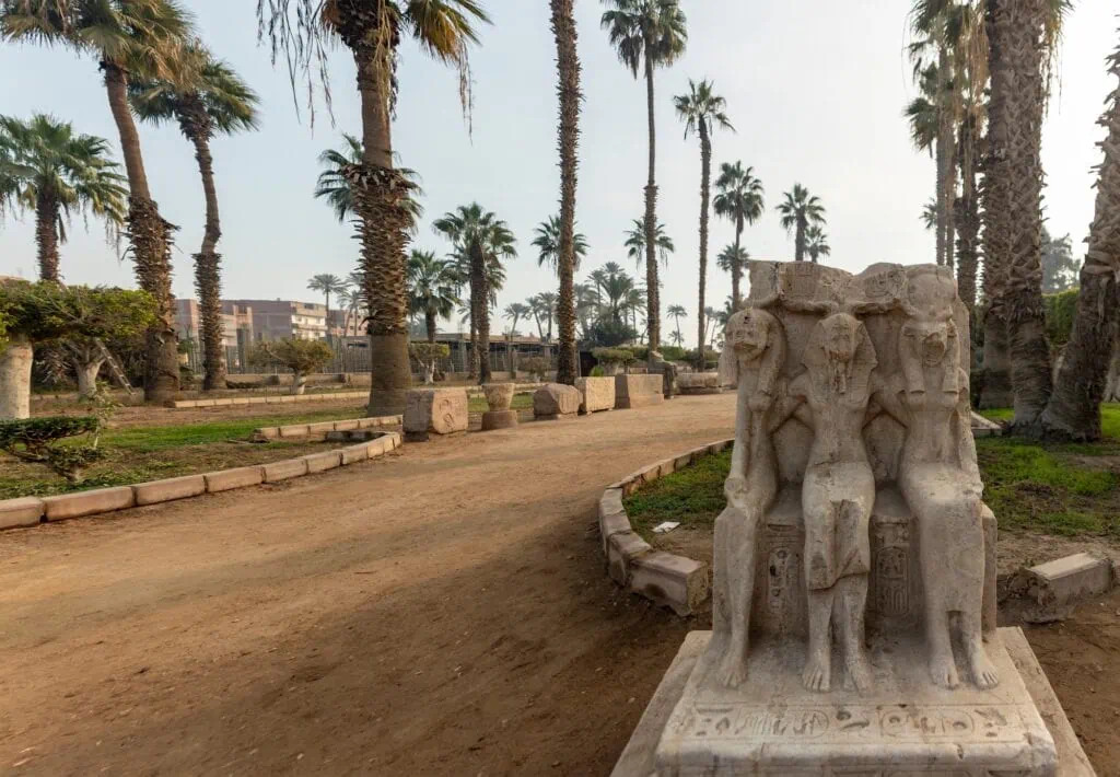 An alabaster triad statue showing three standing figures displayed among ruins at the ancient site of Memphis, Memphis