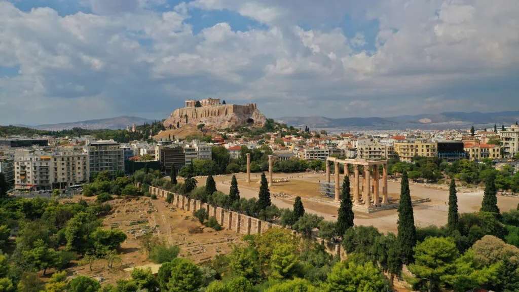 View of the Temple of Olympian Zeus (Olympieion) in Athens, Greece, with the Parthenon on the Acropolis visible on the hillside behind — the largest temple in classical Greece, finally completed under Roman Emperor Hadrian in 131 AD after 700 years of intermittent construction