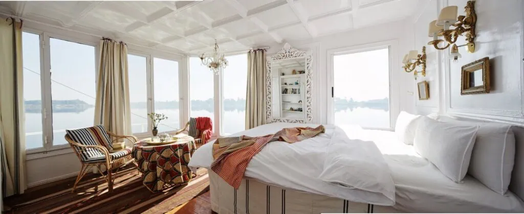 The interior of a panoramic suite cabin aboard a traditional Egyptian dahabiya, with a seating area, large picture windows opening to the Nile, and wooden detailing matching the vessel's traditional character.
