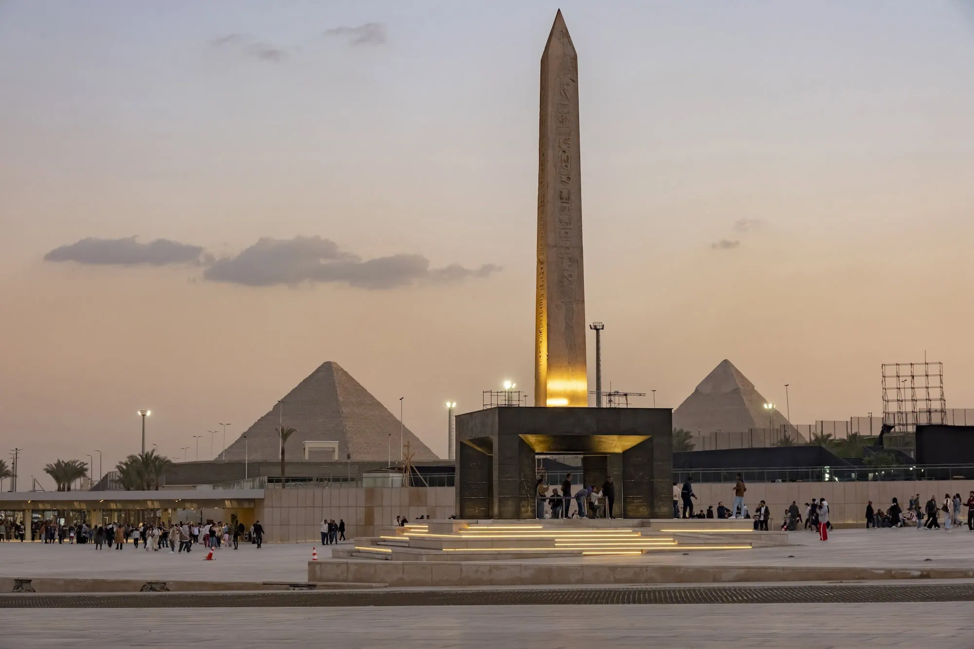 The Grand Egyptian Museum at dusk in Giza, with the illuminated hanging obelisk of Ramses II at the entrance and the silhouettes of the Pyramids of Giza visible in the background