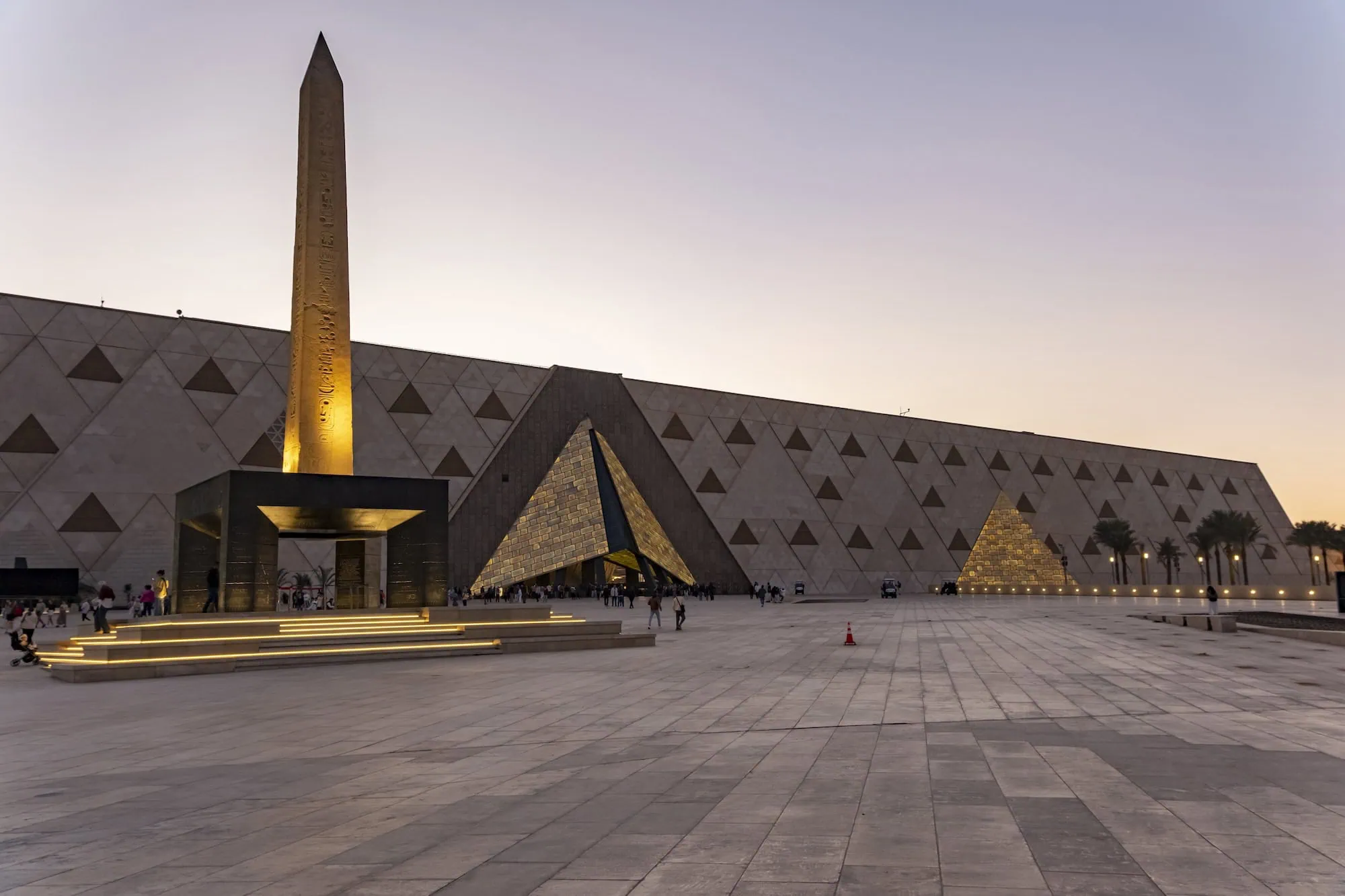 Evening view of the Grand Egyptian Museum's modern triangular architecture with illuminated obelisk and visitors