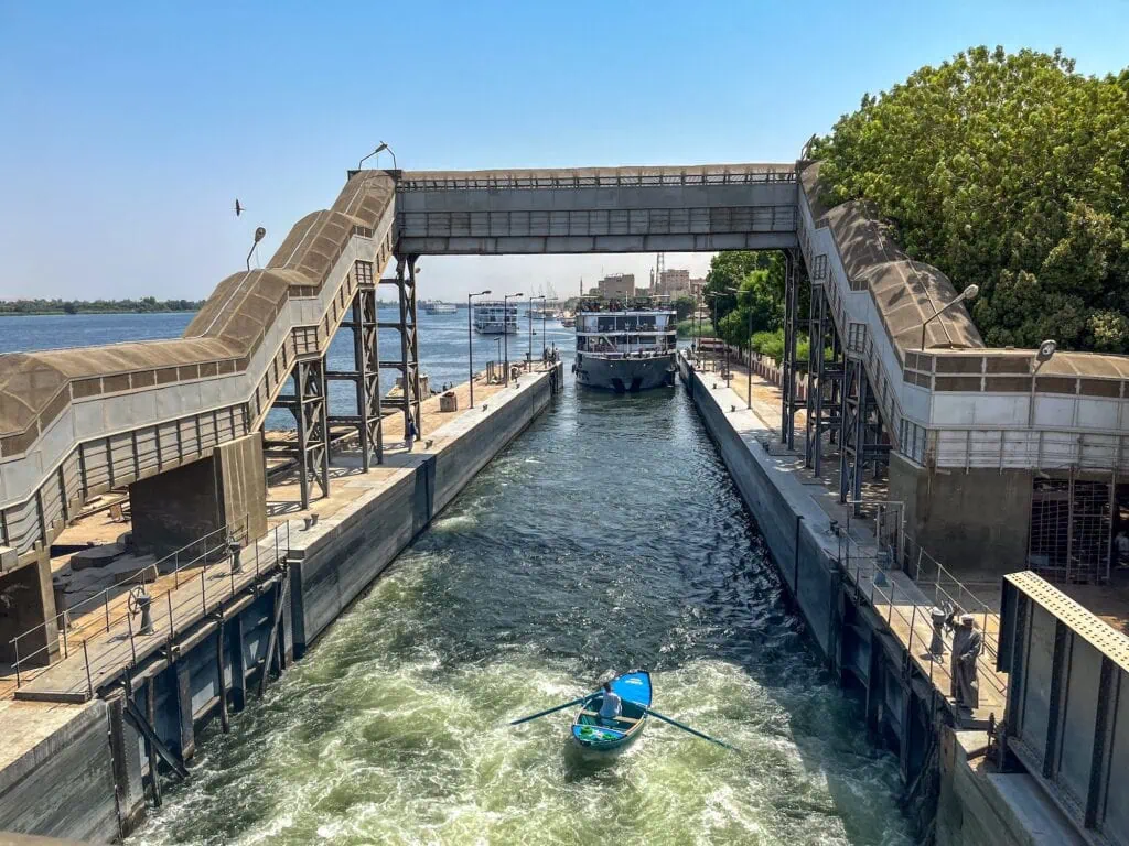 A Nile cruise ship visible at the far end of the Esna Lock chamber after passing through, with a small local rowing boat in the foreground moving through the open lock gates, with the lock infrastructure and pedestrian bridge framing the scene.
