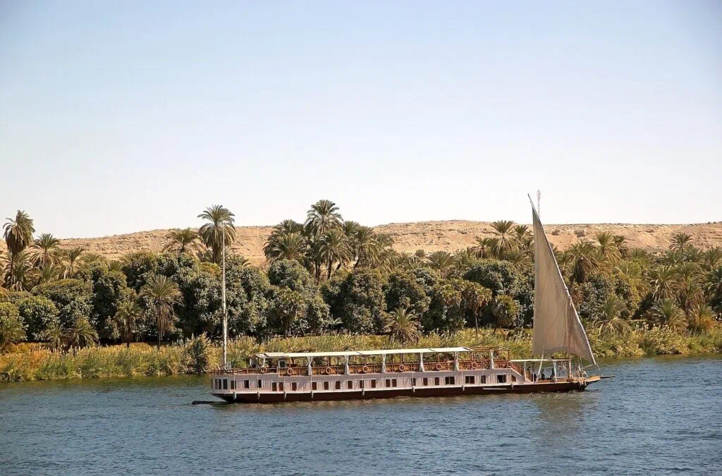 A traditional Egyptian dahabiya with two masts moored along the Nile River with palm trees lining the riverbank