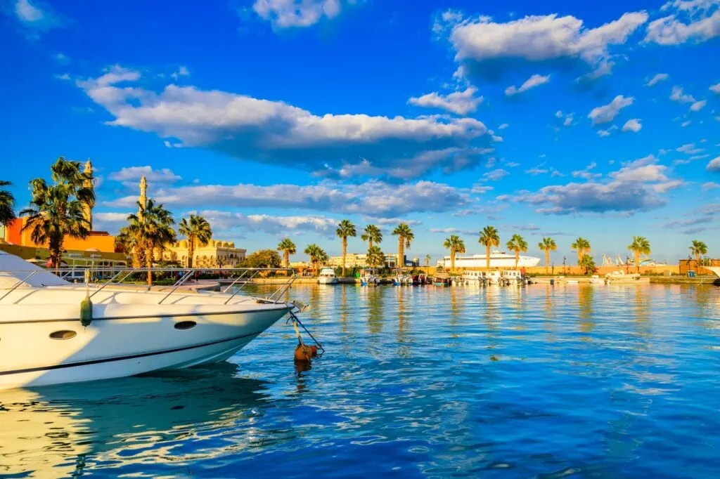 A view of Hurghada Marina on the Red Sea coast of Egypt, with white luxury yachts and tourist boats moored along the harbor edge, with the turquoise Red Sea visible in the background.
