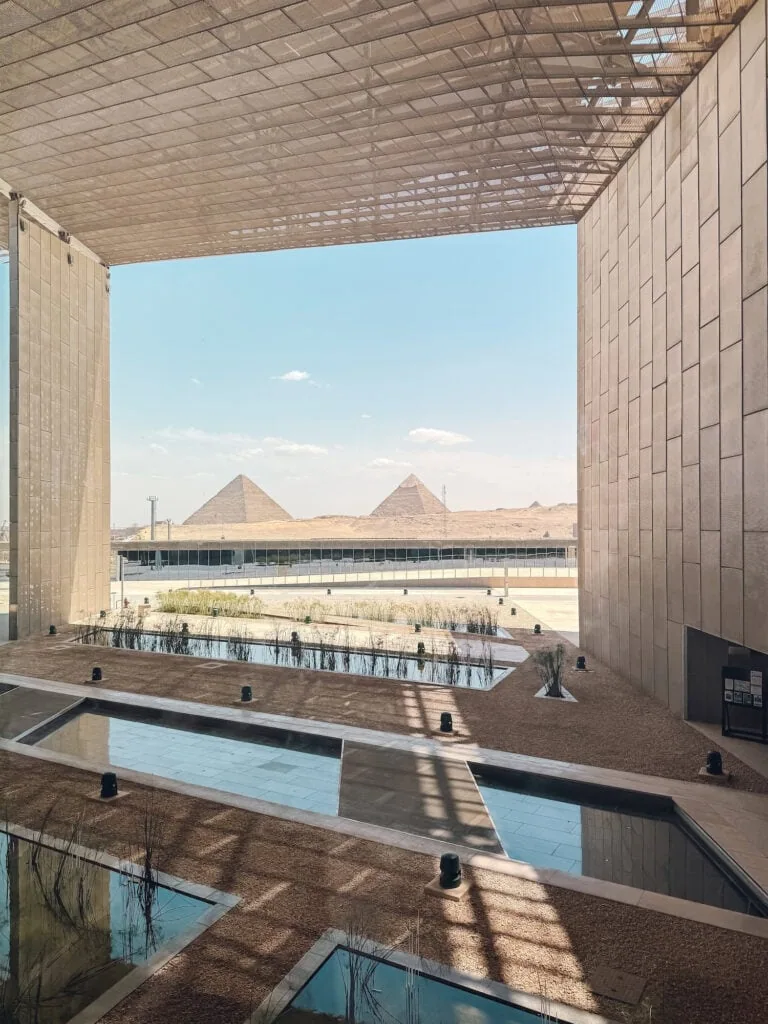 The Pyramids of Giza seen through the large glass facade of the Grand Egyptian Museum, with the museum's interior in the foreground and the pyramids rising in the distance.