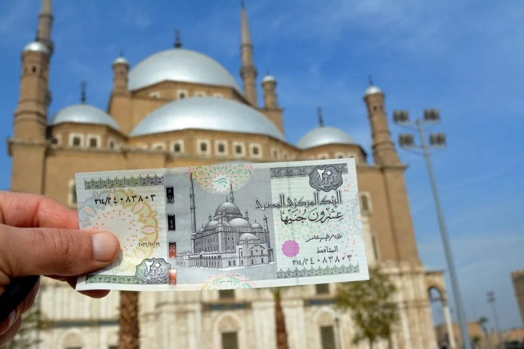 Egyptian 20-pound banknote held up against the Mosque of Muhammad Ali in Cairo, the same Citadel landmark depicted on the note's reverse side.
