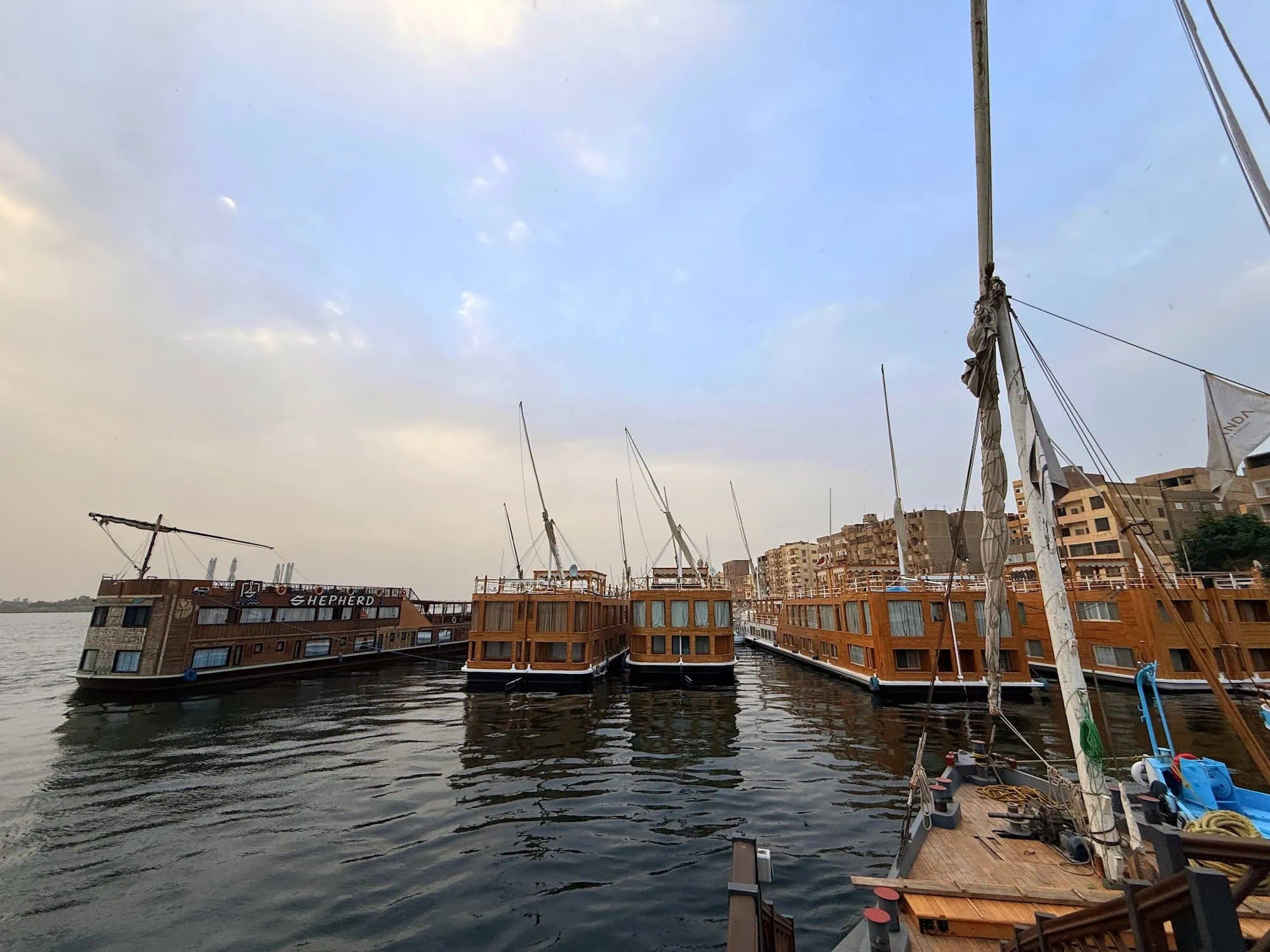 Multiple traditional wooden Egyptian dahabiya sailing vessels moored side by side along the Nile River, with their twin masts visible and lateen sails furled, photographed in clear afternoon light.