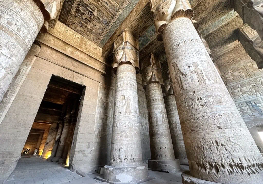 Massive hieroglyph-covered columns and a stone inner doorway inside the Temple of Hathor at Dendera, photographed in low interior light filtering from above.