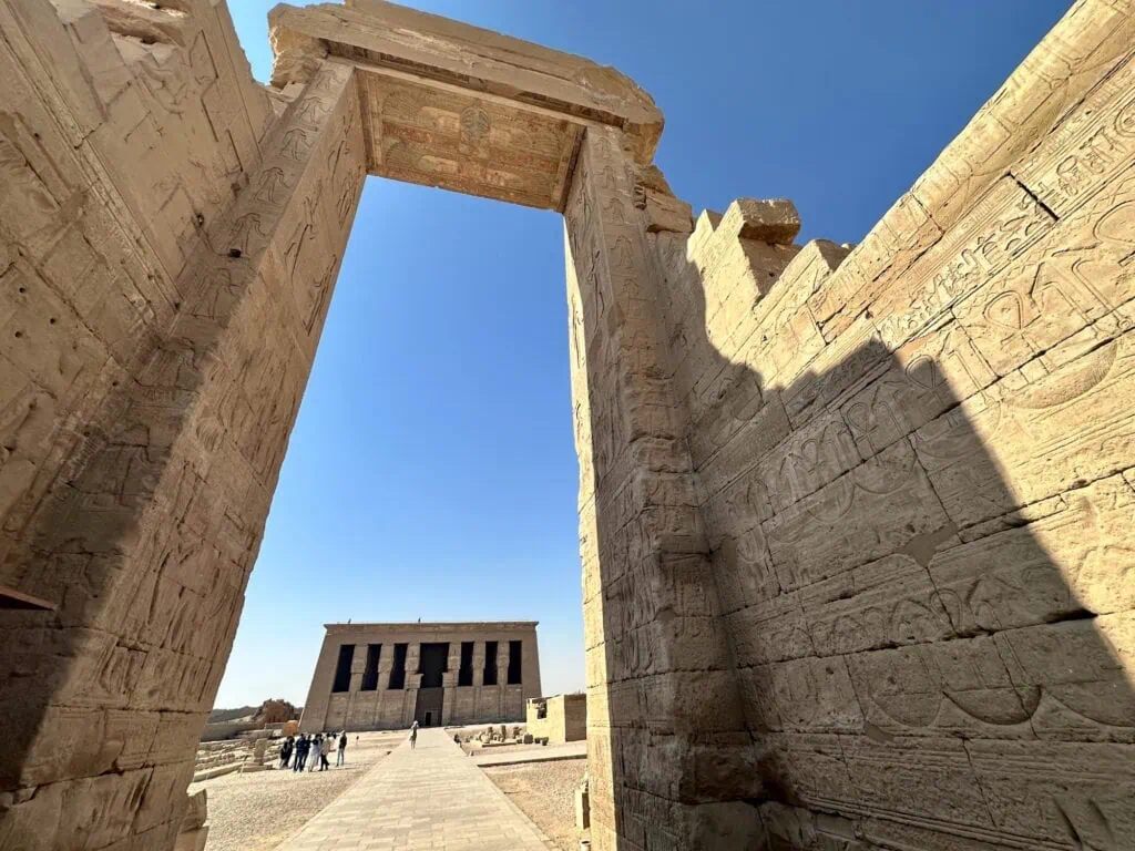 The Hathor temple at Dendera seen through a massive stone gateway carved with hieroglyphic reliefs, with the temple's columned facade visible in the distance beyond the portal.