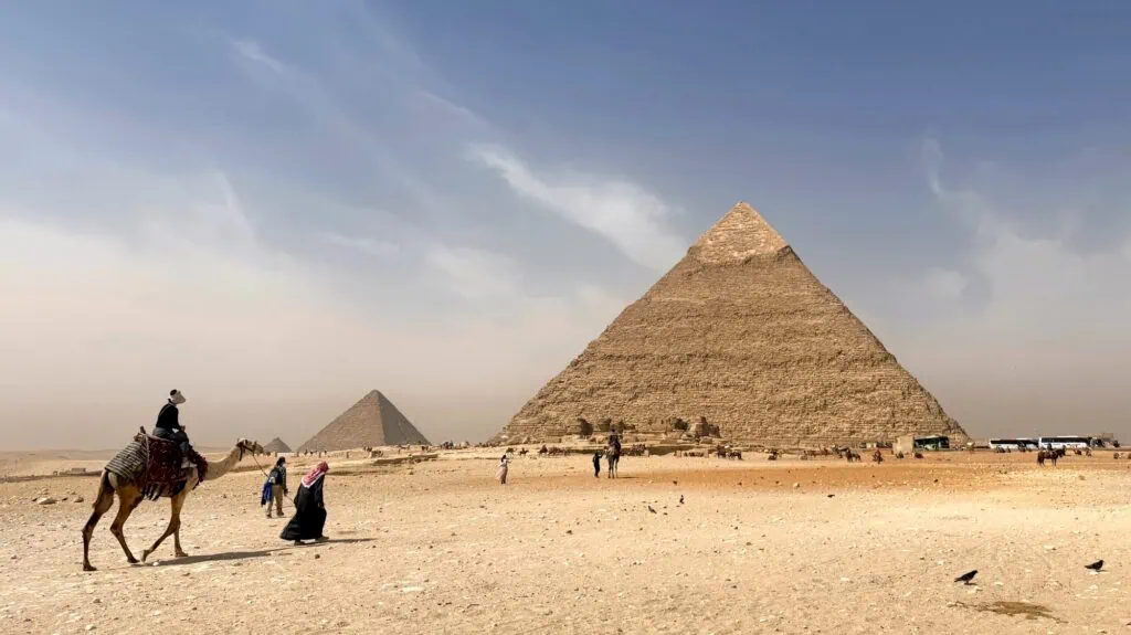 A tourist riding a camel on the Giza Plateau with the Pyramids of Giza rising in the background under a clear sky near Cairo.