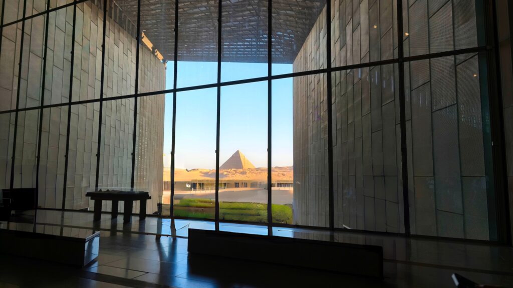 View of the Pyramids of Giza framed by the panoramic windows of the Grand Egyptian Museum (GEM) in Giza, Egypt — the museum's facade is positioned on a direct sightline to the Pyramids, two kilometres away on the same plateau