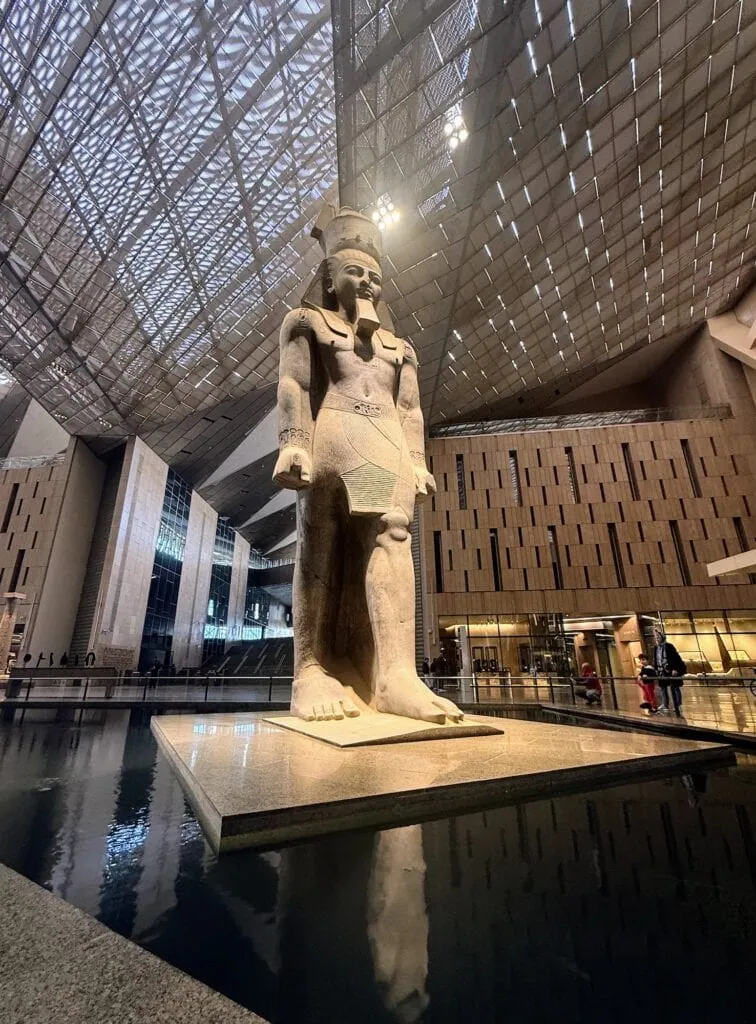 The Grand Hall (Grand Staircase) of the Grand Egyptian Museum (GEM) in Giza, Egypt, with the colossal granite statue of Ramses II at its centre — 11 metres tall, 83 tonnes, the museum's signature object on the central axis