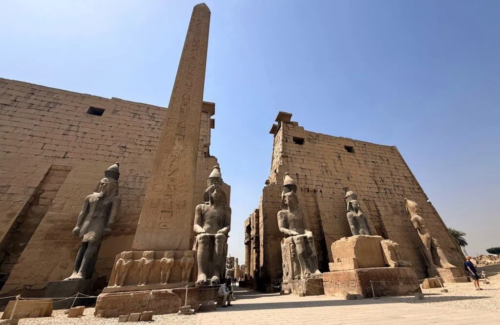 The pylon entrance of Luxor Temple with seated colossal statues of Ramses II flanking the gate and one surviving red granite obelisk in front, photographed in late-afternoon sun. 5-Day Nile Cruises