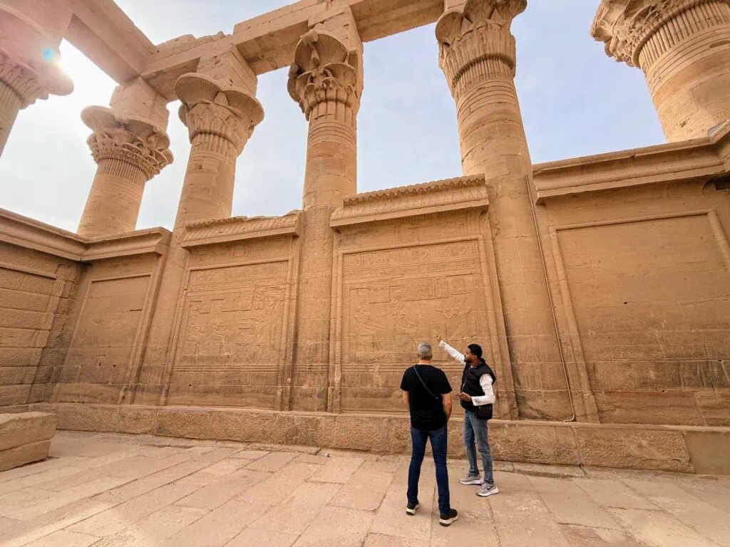A licensed Egyptologist guide explaining the architecture of Trajan's Kiosk at Philae Temple complex in Aswan to a cruise traveler, with the kiosk's columns and architraves visible behind.